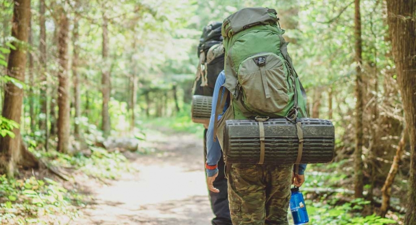 A person wearing a large backpack holds a water bottle as they hike away from the camera on a dirt trail lined with green trees. 